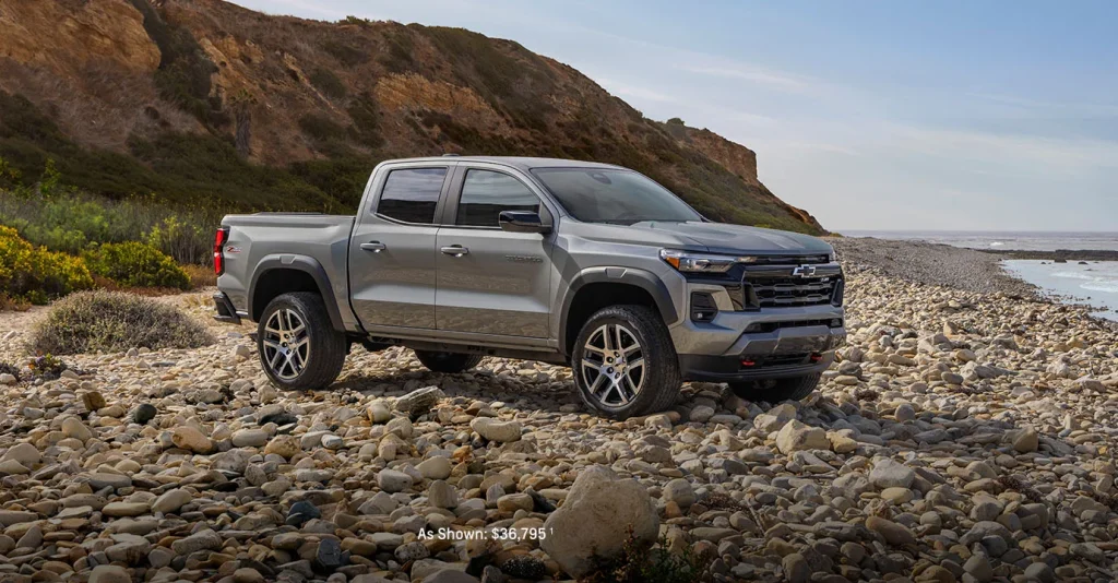 A silver Chevy Colorado parked on rocky terrain by the shore, near Mangino Chevrolet.