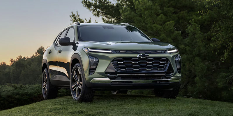 A green 2025 Chevy Trax parked on a grassy hill in a scenic park at sunset near Amsterdam, NY, with warm sunlight reflecting off the compact SUV.