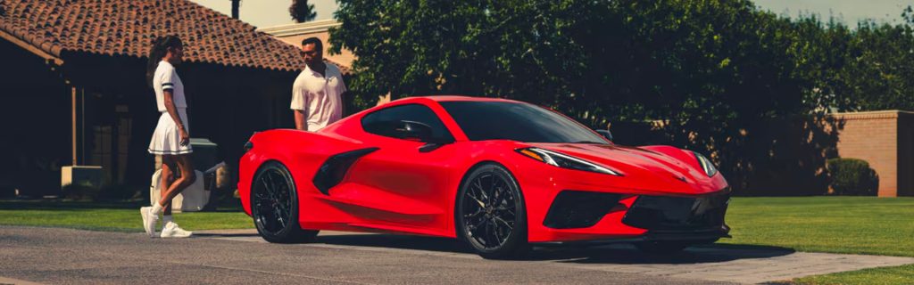 Red Chevrolet Corvette sports car parked in front of a modern house, with a man and woman standing nearby enjoying the view of the luxury performance vehicle.