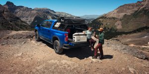 2025 Chevrolet Silverado 1500 in blue parked on a mountain overlook near Amsterdam, NY, with two people enjoying an outdoor adventure, showcasing the truck’s off-road capability and versatility.