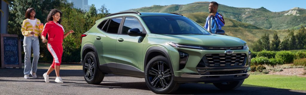 Front three-quarter view of a green 2025 Chevrolet Trax parked outdoors with three people walking nearby in a scenic hillside setting.