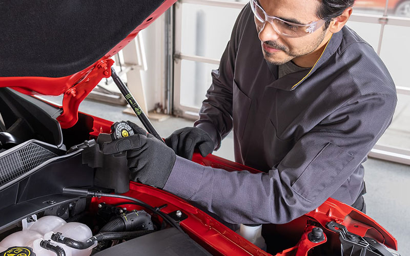 Auto technician wearing safety gloves and glasses inspecting a vehicle engine under an open hood inside a service garage.