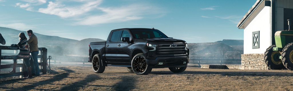 Exterior of a black Chevy Silverado 1500 truck showing the front and passenger side, parked at a farm with mountains in the background.
