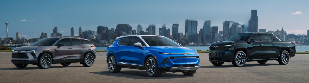 A lineup of Chevrolet electric vehicles, including a silver Chevy Equinox EV, a blue Chevy Blazer EV, and a black Chevy Silverado EV, set against a city skyline in the background.