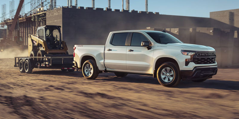 Side view of a Chevrolet pickup truck towing a construction skid-steer loader on a trailer through a dusty construction site with buildings in the background.