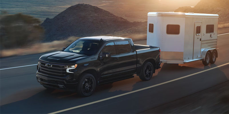 Black Chevrolet Silverado pickup truck towing a travel trailer on a scenic highway at sunset