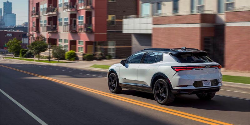 A white 2025 Chevrolet Equinox EV driving toward a downtown cityscape.