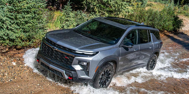 Gray Chevrolet Traverse driving through a shallow stream surrounded by trees.
